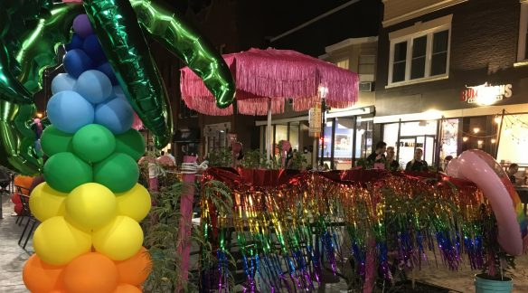 An evening photo of Division Street with rainbow streamers, a pink umbrella, and an inflatable rainbow palm tree.